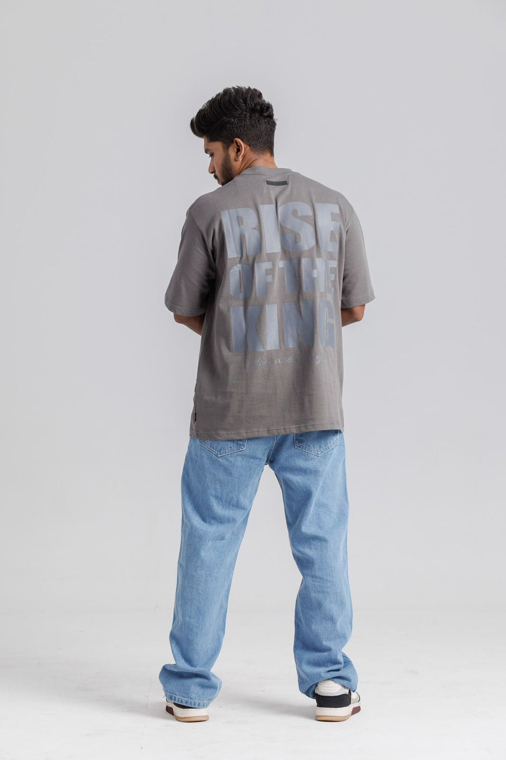 Person wearing a gray t-shirt with text on the back and blue jeans, standing against a white background