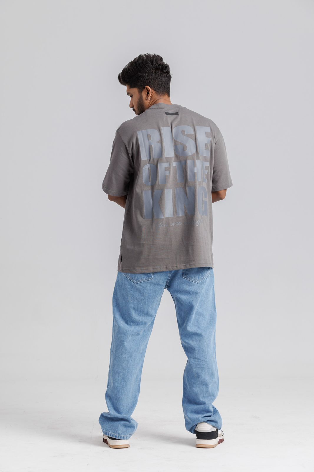 Person wearing a gray t-shirt with text on the back and blue jeans, standing against a white background