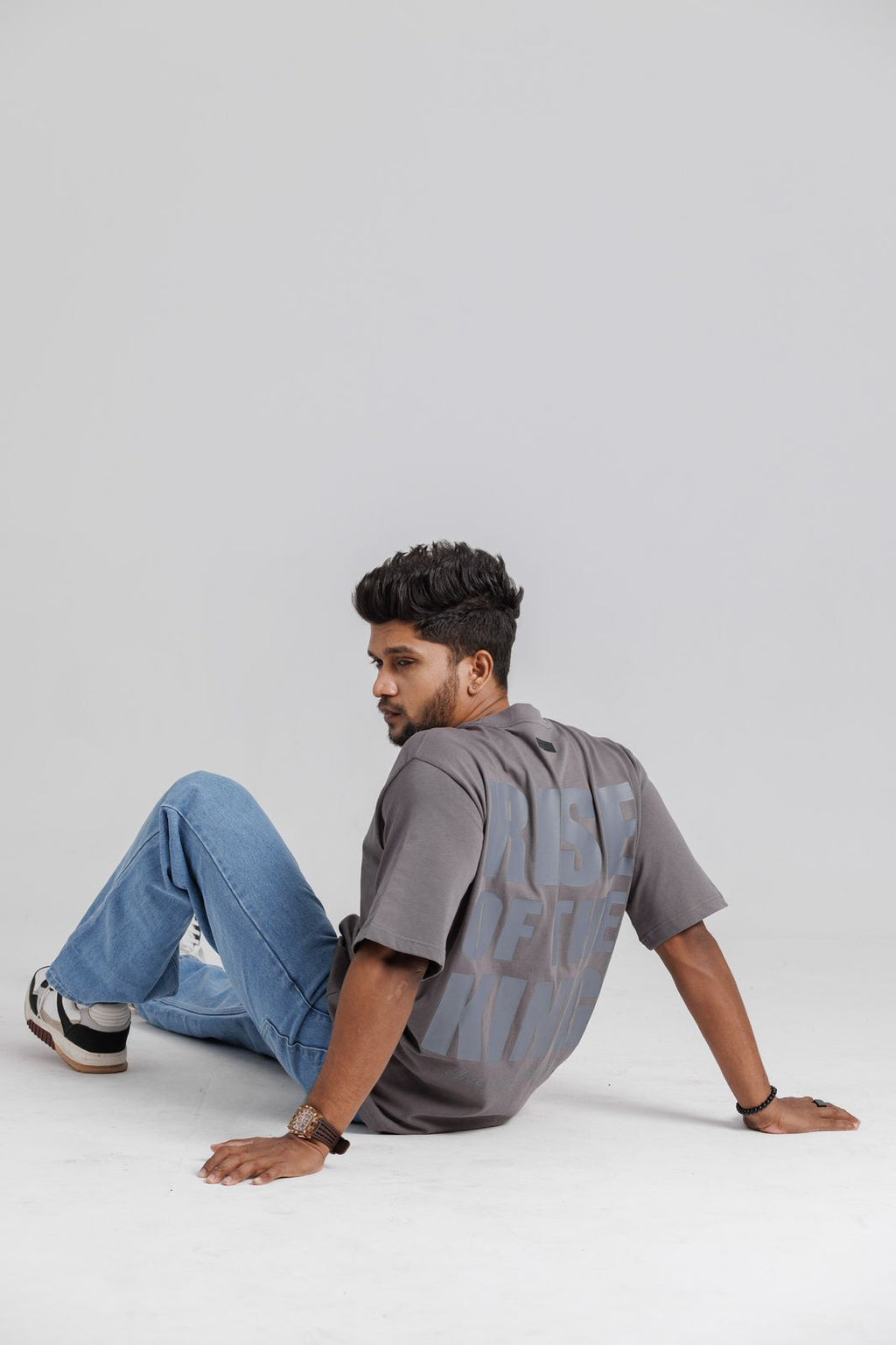 Man sitting on a white floor wearing a gray shirt and blue jeans against a light gray background