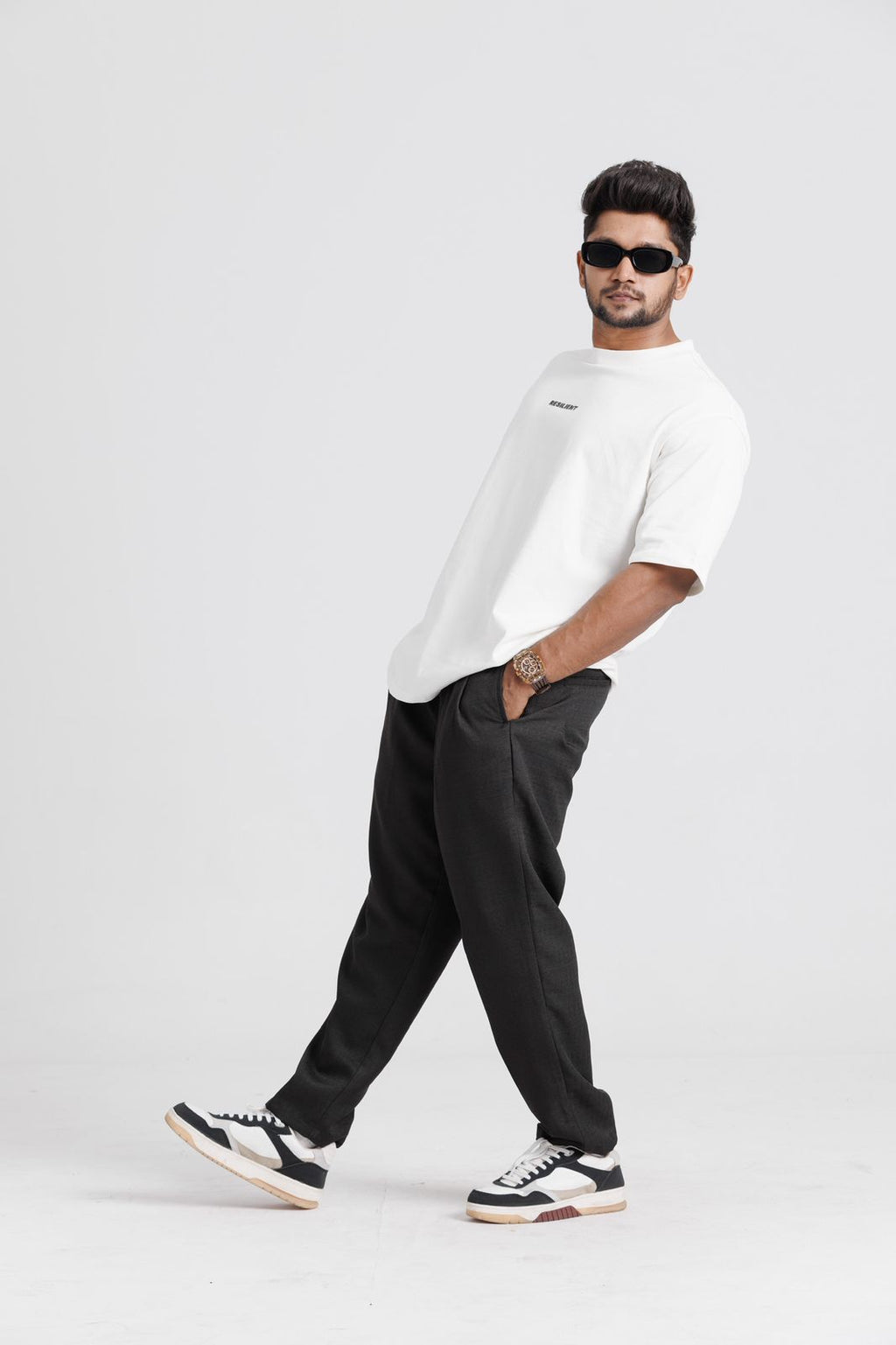 Man wearing a white oversized-shirt, black pants, and black and white sneakers on a light gray background