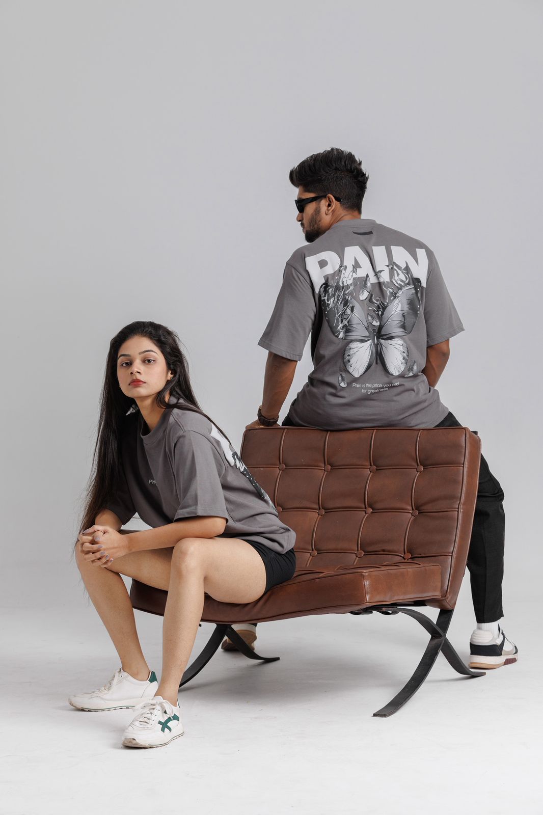 Two people sitting on a brown leather chair wearing gray t-shirts with 'PAIN' text.